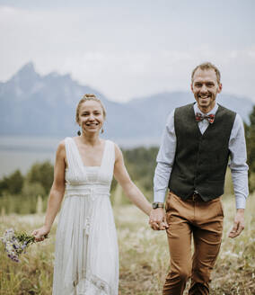 Glücklich lächelnde frisch verheiratete Braut und Bräutigam gehen durch Feld in Tetons - CAVF74660