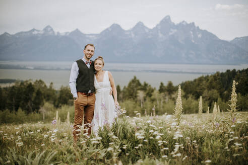 Braut und Bräutigam lächeln und umarmen sich in einem Feld mit Wildblumen - CAVF74667