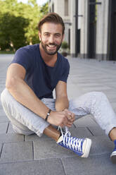 Porträt eines lächelnden Mannes, der in der Stadt auf dem Boden sitzt und seine Schuhe schnürt - PNEF02231