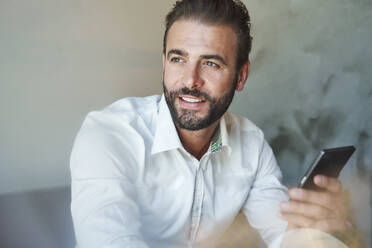 Porträt eines lächelnden Geschäftsmannes mit weißem Hemd, der ein Mobiltelefon hält - PNEF02300