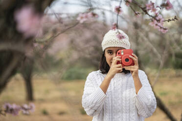 Porträt eines jungen Mädchens, das inmitten blühender Mandelbäume mit der Kamera fotografiert - DLTSF00470