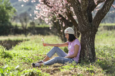 Glückliches Teenager-Mädchen lehnt an einem Mandelbaum und betrachtet einen Zweig mit Mandelblüten - DLTSF00472