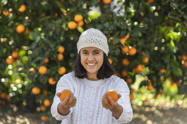 Portrait of happy teenage girl in front of orange tree offering oranges - DLTSF00476