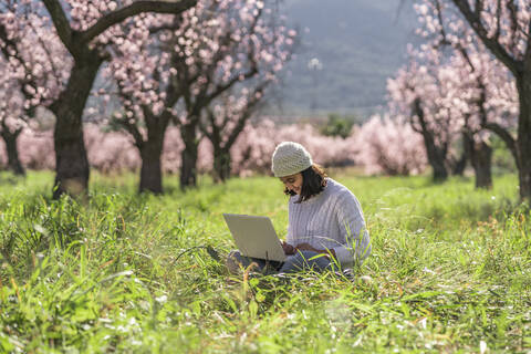 Teenager-Mädchen sitzt auf einer Wiese inmitten blühender Mandelbäume und benutzt einen Laptop, lizenzfreies Stockfoto