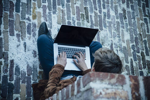 Teenager using laptop and sitting on a stone floor in the city - ANHF00195