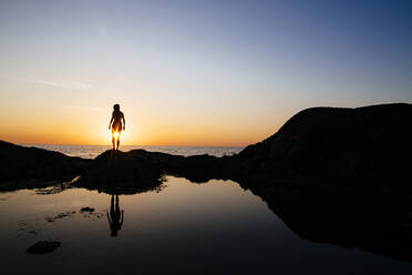 Silhouette einer Frau, die bei Sonnenuntergang auf einem Felsen am Meer steht. - MINF13452