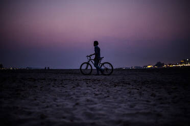 Silhouette eines Jungen, der ein Fahrrad am Ufer eines Flusses bei Sonnenuntergang schiebt. - MINF13549