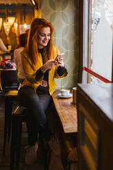 Porträt einer glücklichen rothaarigen jungen Frau, die in einem Café sitzt und ein Handy benutzt - LJF01273