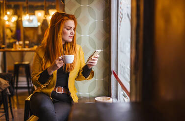 Rothaarige junge Frau sitzt in einem Café mit einer Tasse Kaffee und schaut auf ihr Handy - LJF01276