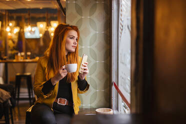 Portrait of redheaded young woman with smartphone waiting in a coffee shop looking out of window - LJF01277