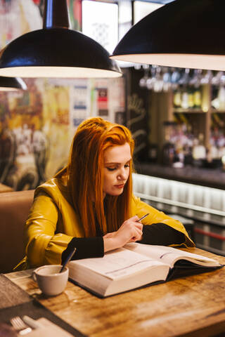 Rothaarige junge Frau am Tisch in einer Kneipe mit Blick auf ein Buch, lizenzfreies Stockfoto
