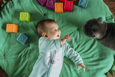 Happy baby girl lying on green mat watching cat - GEMF03421