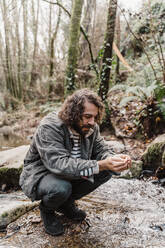 Young man crouching at stream in forest, drinking water from cupped hands - AFVF05271