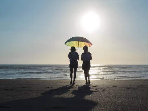 Zwei Frauen mit Sonnenschirm, die in der Abenddämmerung am Strand spazieren gehen, Strand Kedungu, Bali, Indonesien, lizenzfreies Stockfoto