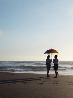 Zwei Frauen mit Sonnenschirm stehen in der Abenddämmerung am Strand, Kedungu Strand, Bali, Indonesien - KNTF04329