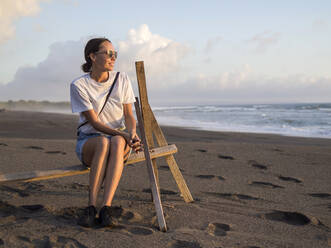 Frau sitzt auf einem Gestell am Strand in der Abenddämmerung, Kedungu Strand, Bali, Indonesien - KNTF04330