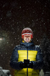 Smiling man using cell phone in snowfall at night - CJMF00249