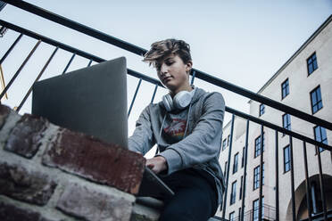 Teenager using laptop in the city, headphones around his neck - ANHF00202