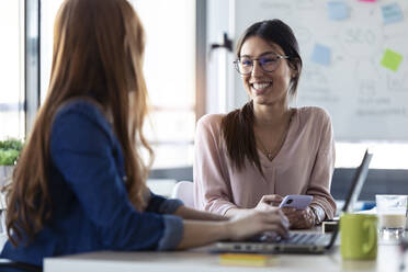 Smiling businesswomen using smartphone, talking with her colleague - JSRF00849