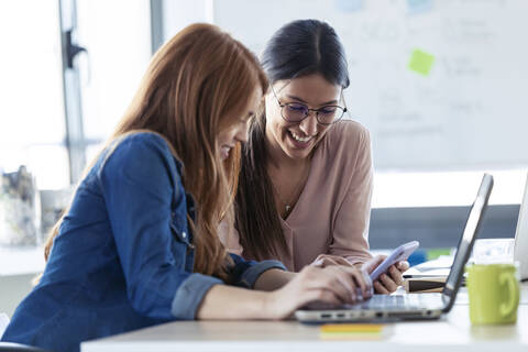 Lächelnde Geschäftsfrauen mit Smartphone und Laptop im Büro, lizenzfreies Stockfoto