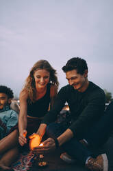 Smiling couple protecting burning candle with hands while sitting by friend on terrace against sky - MASF16638