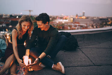 Smiling young couple protecting burning candle with hands while sitting on terrace against sky during dusk - MASF16639