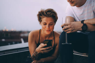 Frau benutzt ihr Smartphone, während sie mit einem Freund auf der Terrasse sitzt und Wein trinkt - MASF16640