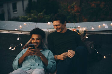 Lächelnde männliche Freunde, die auf der Terrasse sitzend auf ihr Smartphone schauen, während einer Dachterrassenparty in der Stadt - MASF16645