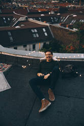 Hoher Winkel in voller Länge eines glücklichen jungen Mannes, der sich auf einer beleuchteten Terrasse während einer Party in der Abenddämmerung entspannt - MASF16646