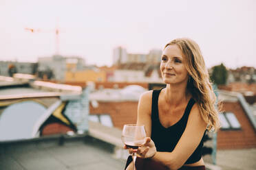 Thoughtful smiling woman having wine while sitting on terrace during rooftop party - MASF16647