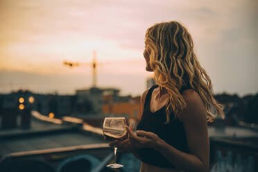 Thoughtful young woman having wine while looking away on terrace during rooftop party at sunset - MASF16648