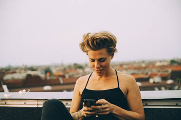 Smiling woman with short hair text messaging on smart phone while sitting on terrace against sky - MASF16651