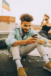 Lächelnder junger Mann, der sein Smartphone benutzt, während er auf der Terrasse einer Party sitzt - MASF16664