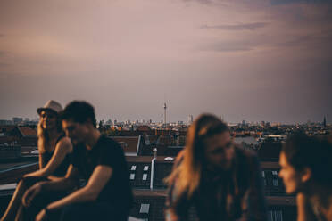 Friends talking while sitting on terrace during rooftop party in city at dusk - MASF16689