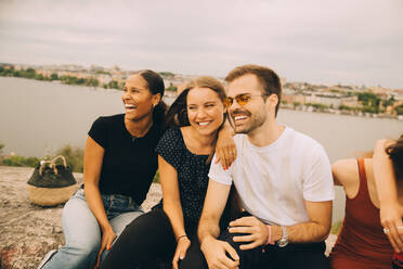 Cheerful friends looking away while sitting together on rock formation against lake - MASF16700