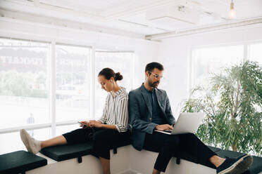 Male and female colleagues using technologies while sitting at corridor in creative office - MASF16720