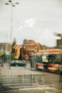 Lächelnder Geschäftsmann, der Kopfhörer trägt und ein Mobiltelefon benutzt, gesehen durch ein Glasfenster im Büro - MASF16738
