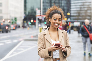 Lächelnde junge Frau mit Handy und Kopfhörern in der Stadt, London, UK - WPEF02554