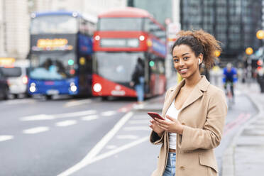 Lächelnde junge Frau mit Handy und Kopfhörern in der Stadt, London, UK - WPEF02556