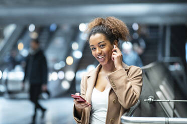Porträt einer glücklichen jungen Frau mit Mobiltelefon und Ohrstöpseln in einer U-Bahn-Station - WPEF02563