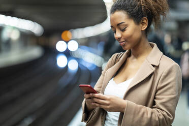 Smiling young woman using cell phone at subway station platform - WPEF02565