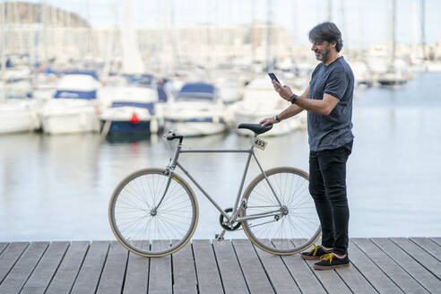 Älterer Mann mit Fixie-Fahrrad steht auf der Promenade und schaut auf sein Handy, Alicante, Spanien - DLTSF00481