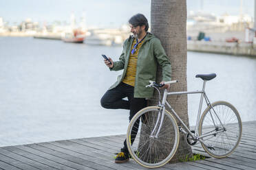 Mature man with fixie bike leaning against palm tree trunk looking at cell phone, Alicante, Spain - DLTSF00493