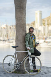 Portrait of relaxed mature man with fixie bike leaning against palm tree trunk, Alicante, Spain - DLTSF00494