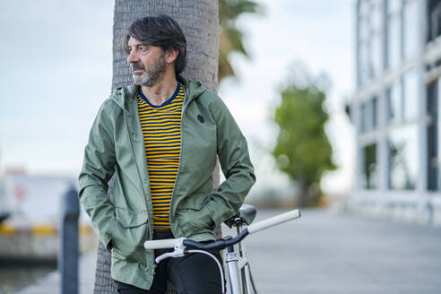 Portrait of serious mature man with bike leaning against palm tree trunk looking at distance, Alicante, Spain - DLTSF00496