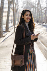 Portrait of smiling young woman with smartphone at the tram stop - AFVF05293