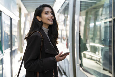 Lächelnde junge Frau mit Smartphone an der Straßenbahnhaltestelle - AFVF05297