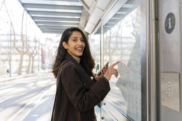 Porträt einer glücklichen jungen Frau, die an der Straßenbahnhaltestelle die Karte kontrolliert - AFVF05305
