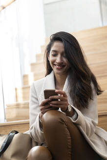 Lächelnde junge Frau sitzt auf einer Treppe und benutzt ein Smartphone - AFVF05319