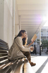 Junge Frau mit Kaffee zum Mitnehmen, die auf einer Bank ein Buch liest - AFVF05347